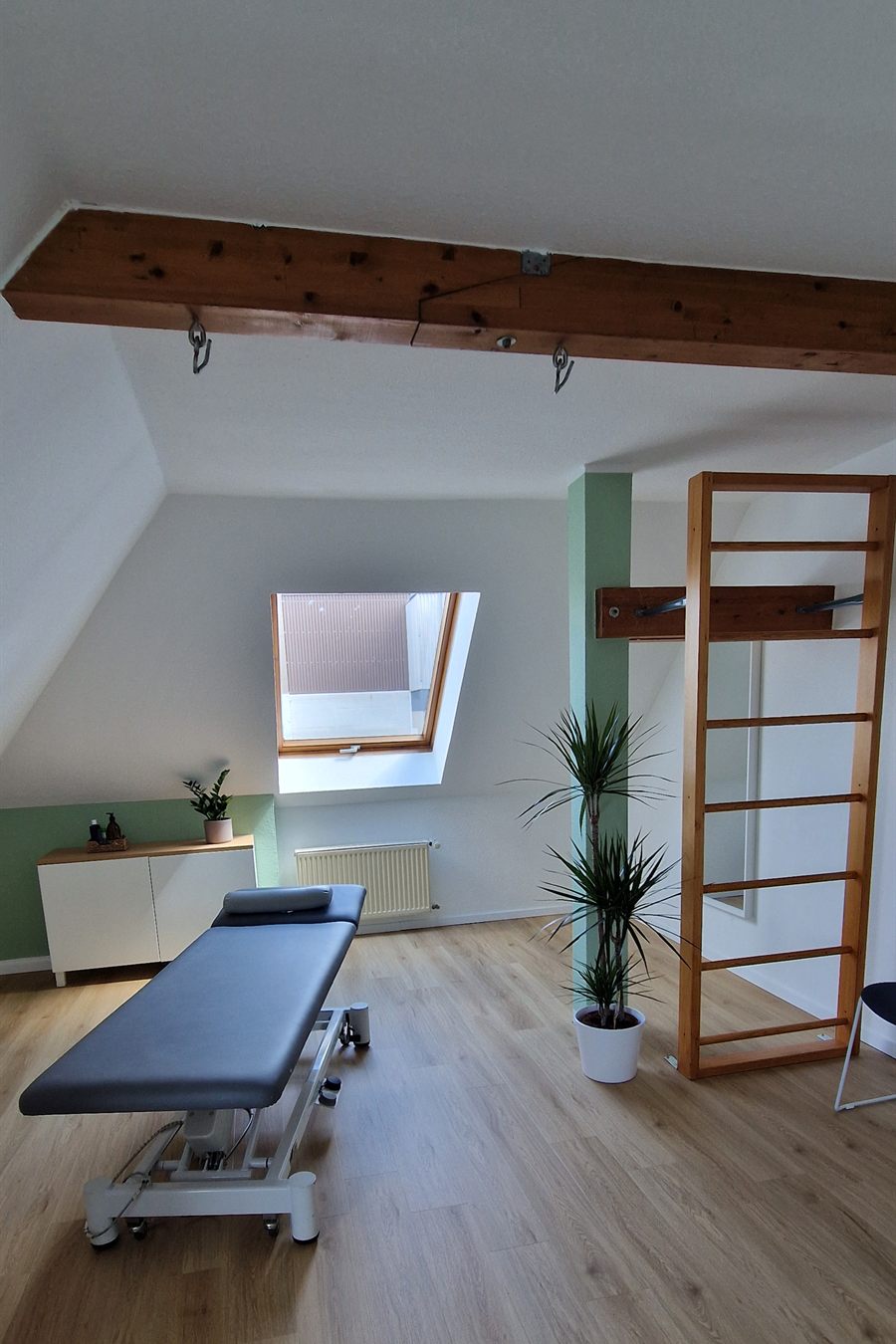 Heller Behandlungsraum unter dem Dach mit Behandlungsliege, Sprossenwand und Dachfenstern.